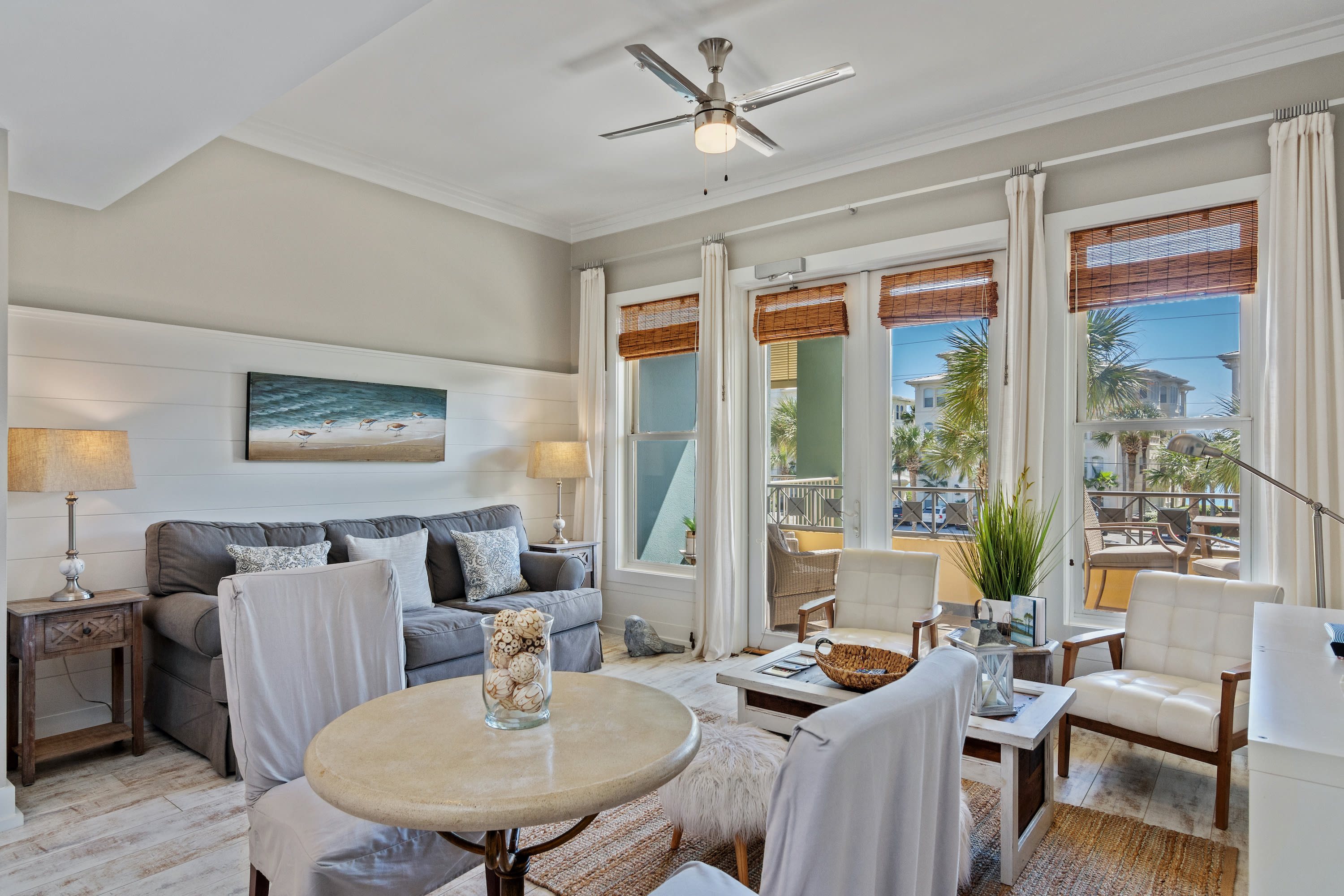 Stylish and sunlit 30A retreat with comfy seating, chic coastal décor, and floor-to-ceiling windows leading to a private balcony with palm views—ideal for relaxing in beachside comfort.
