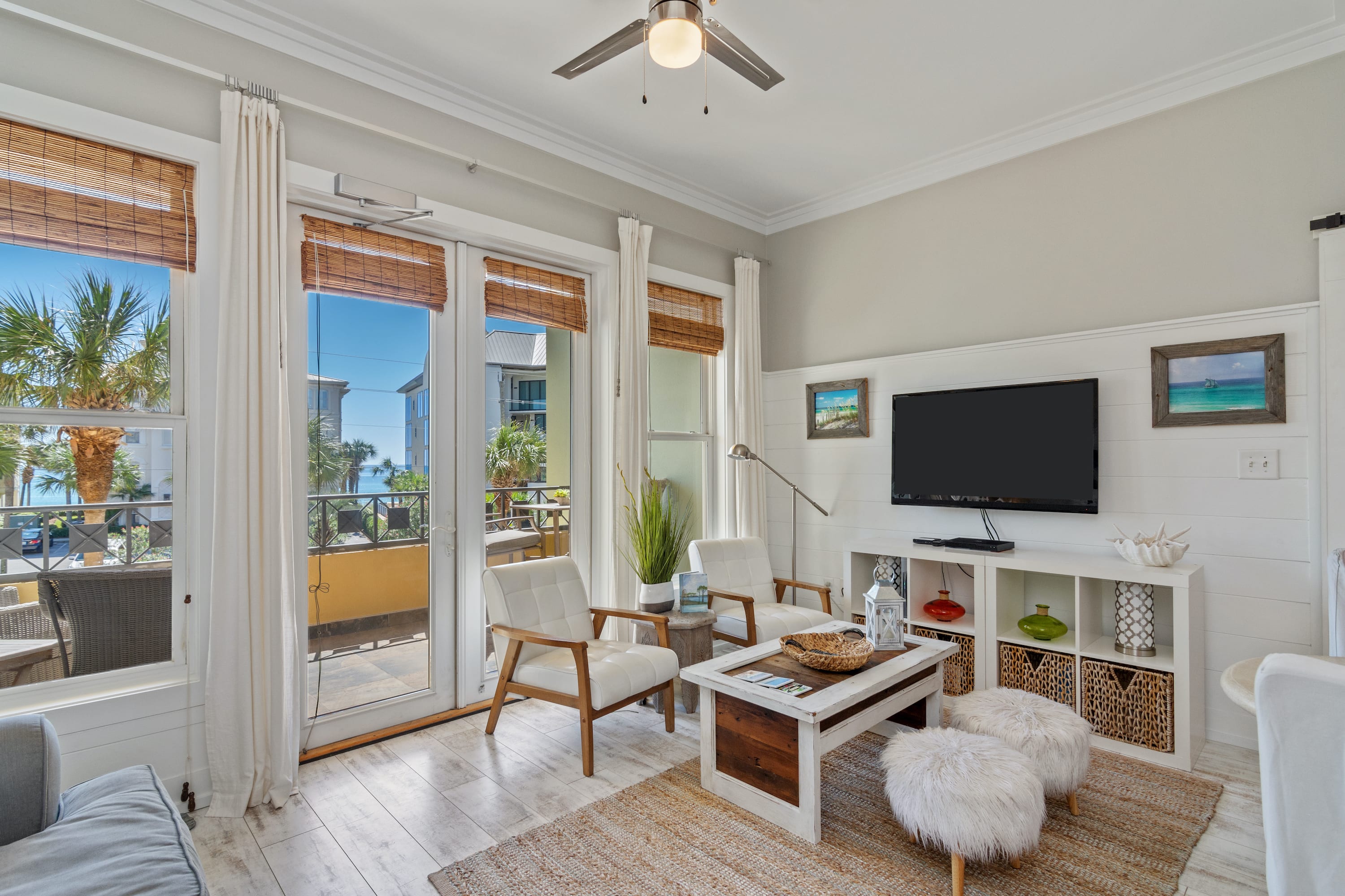 Light-filled living area with coastal flair, featuring comfy seating, a flat-screen TV, and glass doors that open to a palm-lined balcony—perfect for a laid-back 30A getaway.