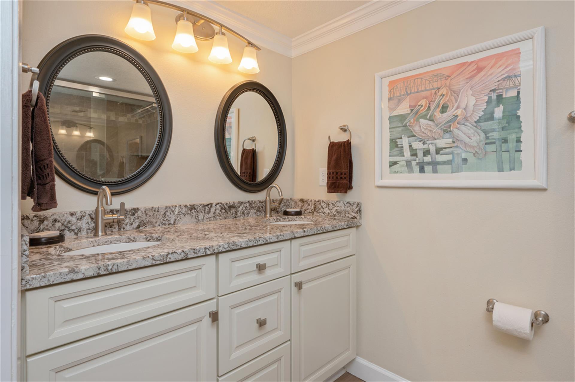 Refresh yourself in this elegant dual-vanity bathroom featuring granite countertops, warm lighting, and thoughtful coastal artwork to brighten your morning routine.