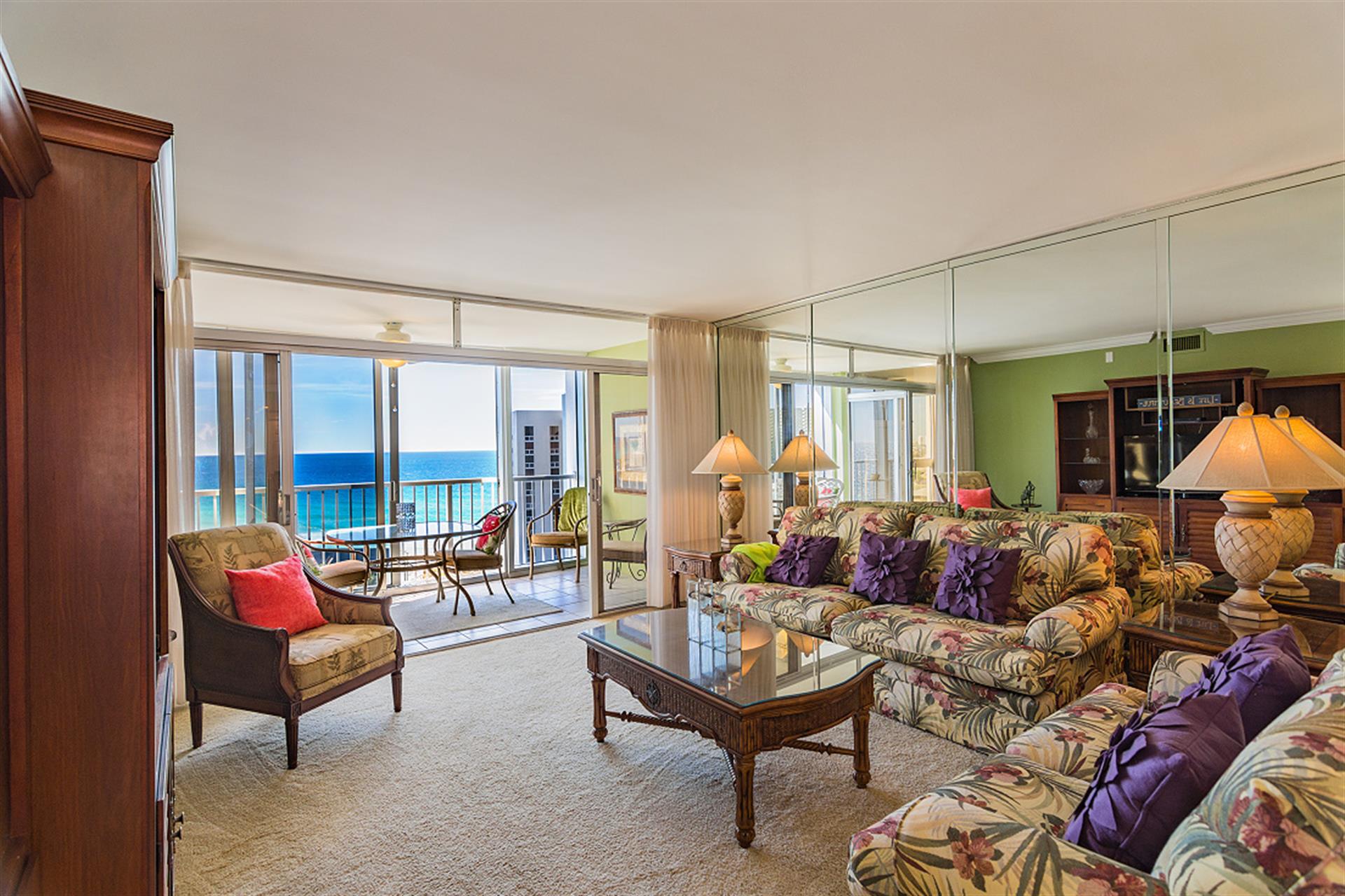 Step into your tropical living room where plush floral seating meets stunning ocean views through floor-to-ceiling windows.