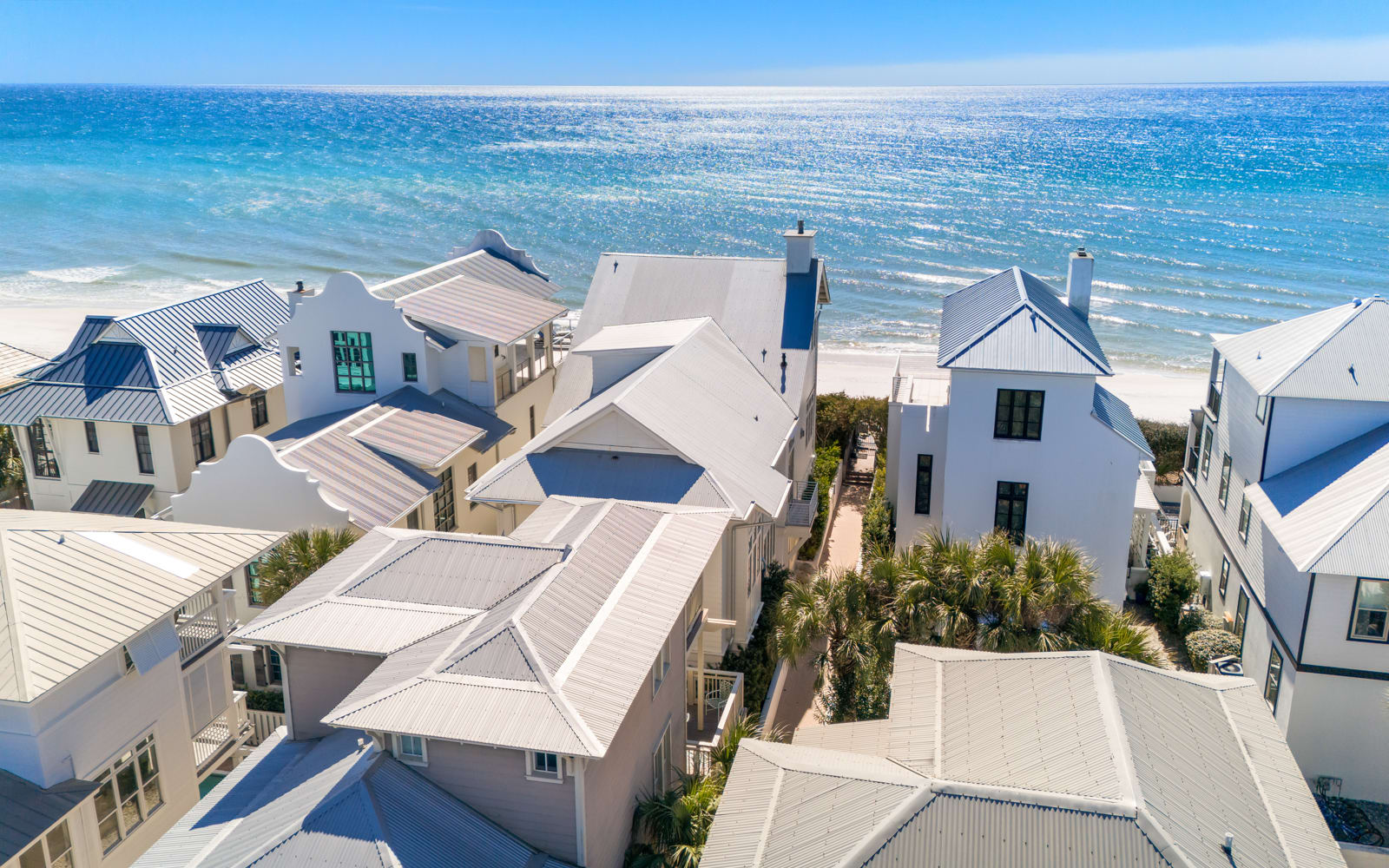 Aerial view of beachfront rental homes featuring white architecture and metal roofs, situated directly on pristine sandy shores with crystal-clear turquoise waters.