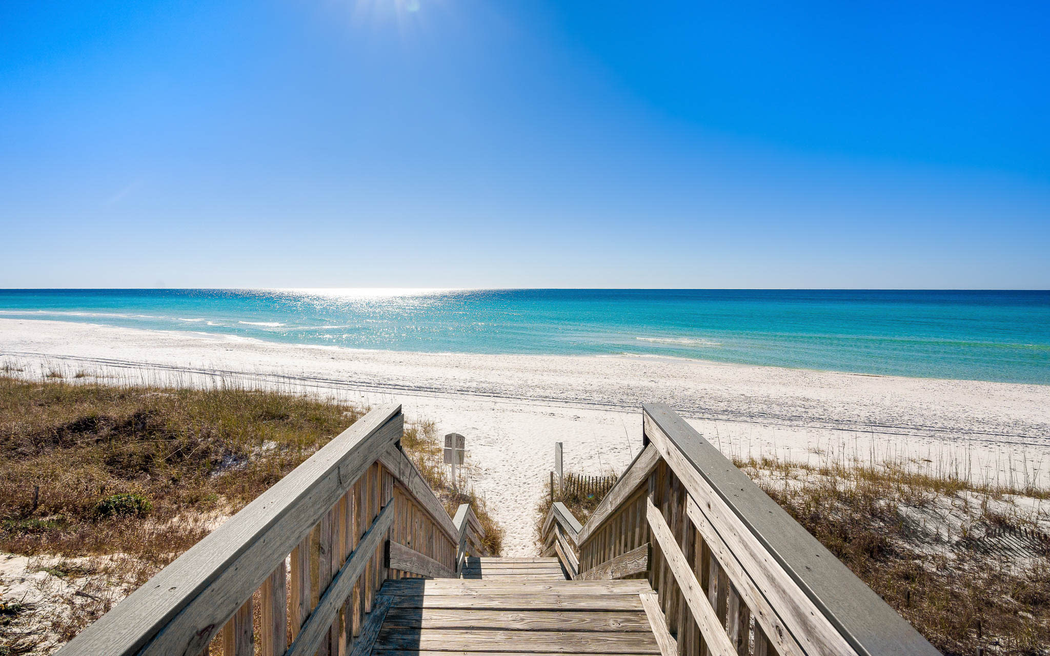 Wooden boardwalk leads to pristine white sand beach with crystal-clear turquoise waters stretching to the horizon.
