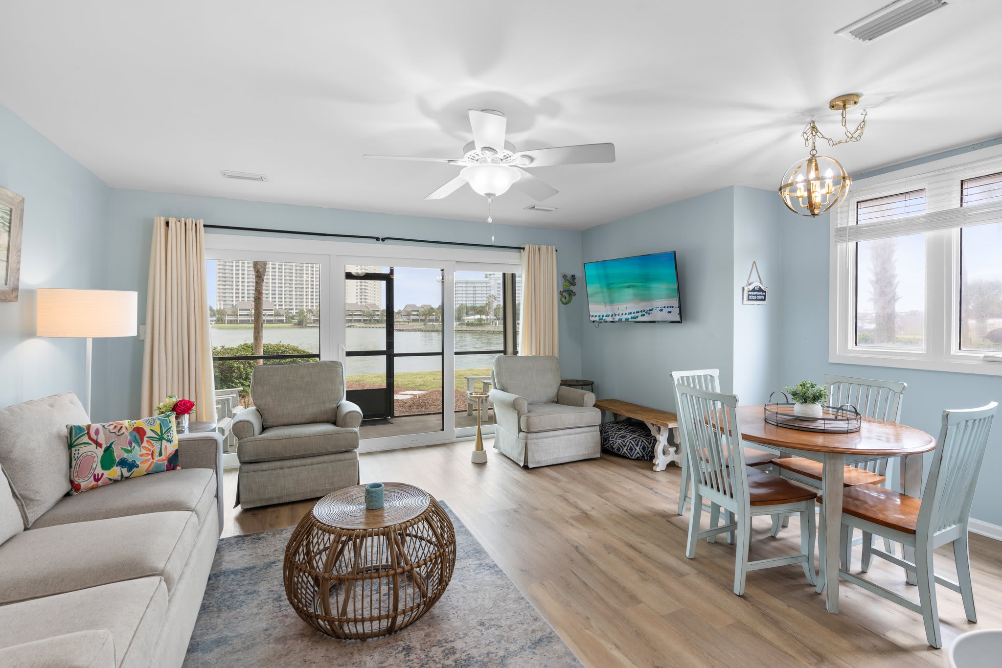 Cozy lakefront living room with armchairs, a rattan coffee table, flat-screen TV, and dining set for four. Sliding glass doors open to serene water views and a private patio.