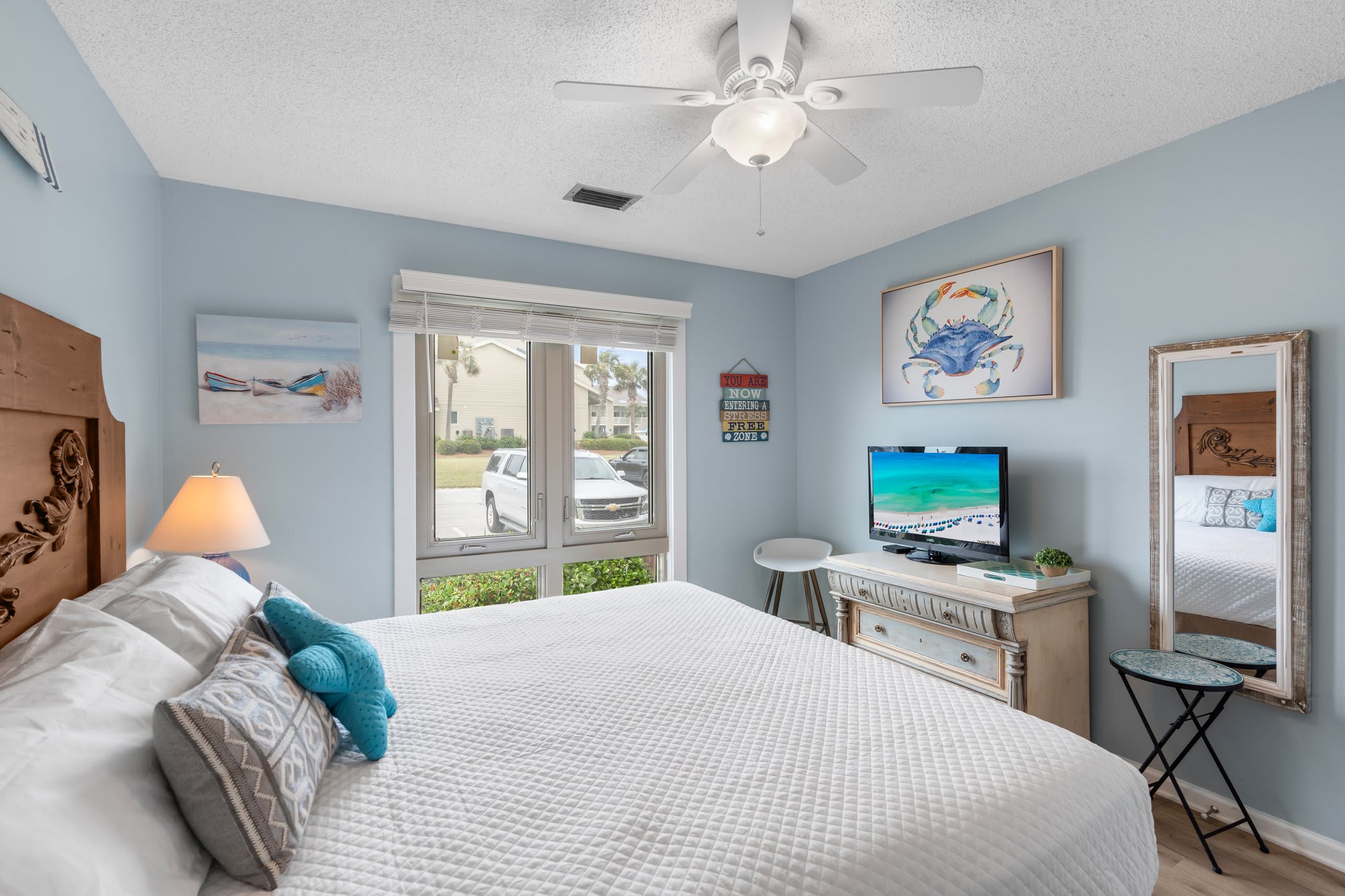 Bright coastal guest bedroom with a comfy king bed, beach-themed decor, flat-screen TV, and natural light pouring in through large windows—your perfect retreat after a day in the sun.