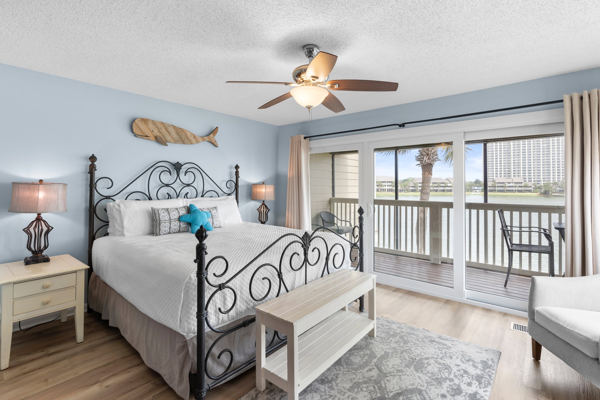 Spacious primary bedroom with a wrought iron king bed, coastal décor, and sliding doors that open to a private balcony with serene lake views—your perfect waterfront retreat.