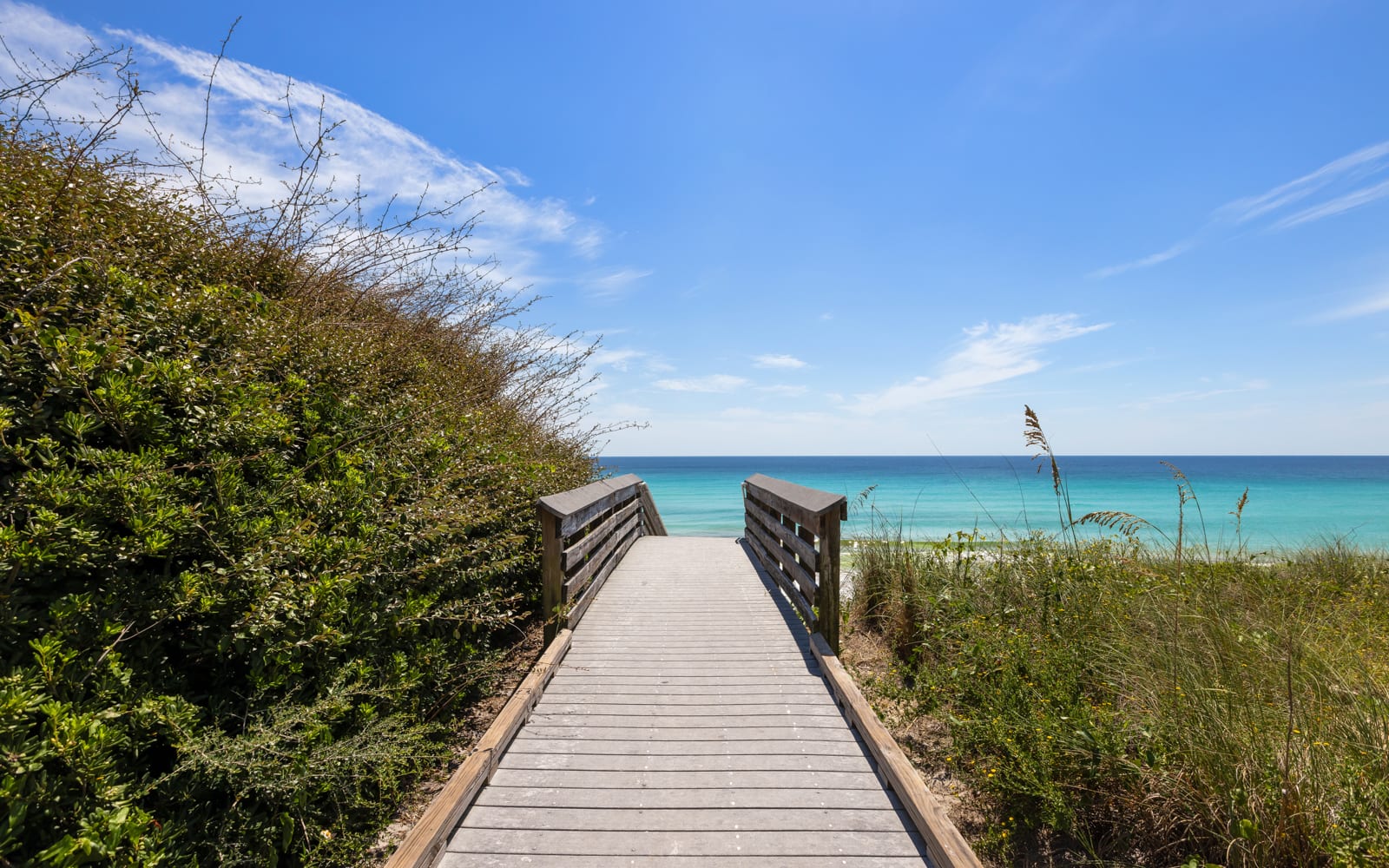Wooden boardwalk leads through coastal vegetation to pristine turquoise waters and sandy beach under brilliant blue skies.