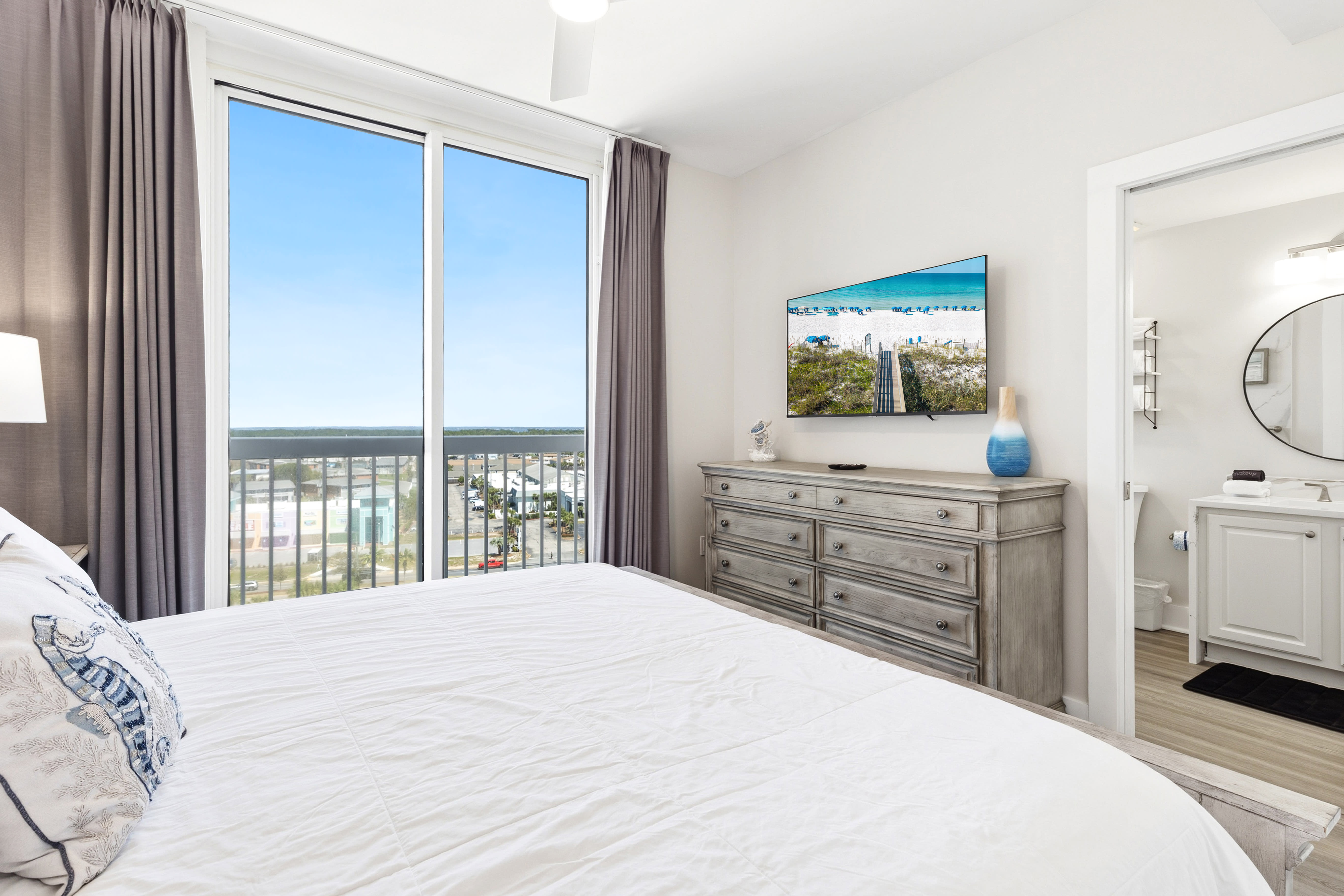 Wake up to stunning ocean views from your private balcony in this bright, elegant bedroom with modern coastal furnishings and TV.