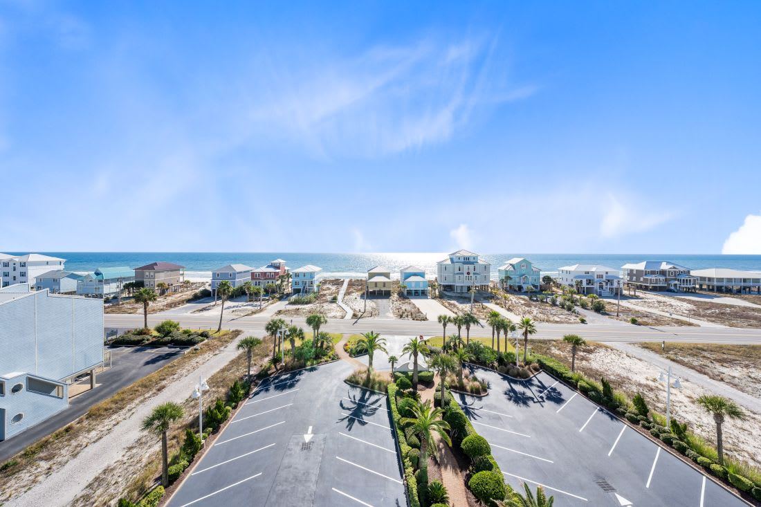 Aerial view of beachfront community with colorful vacation homes and landscaped parking area surrounded by palm trees and white sand dunes.