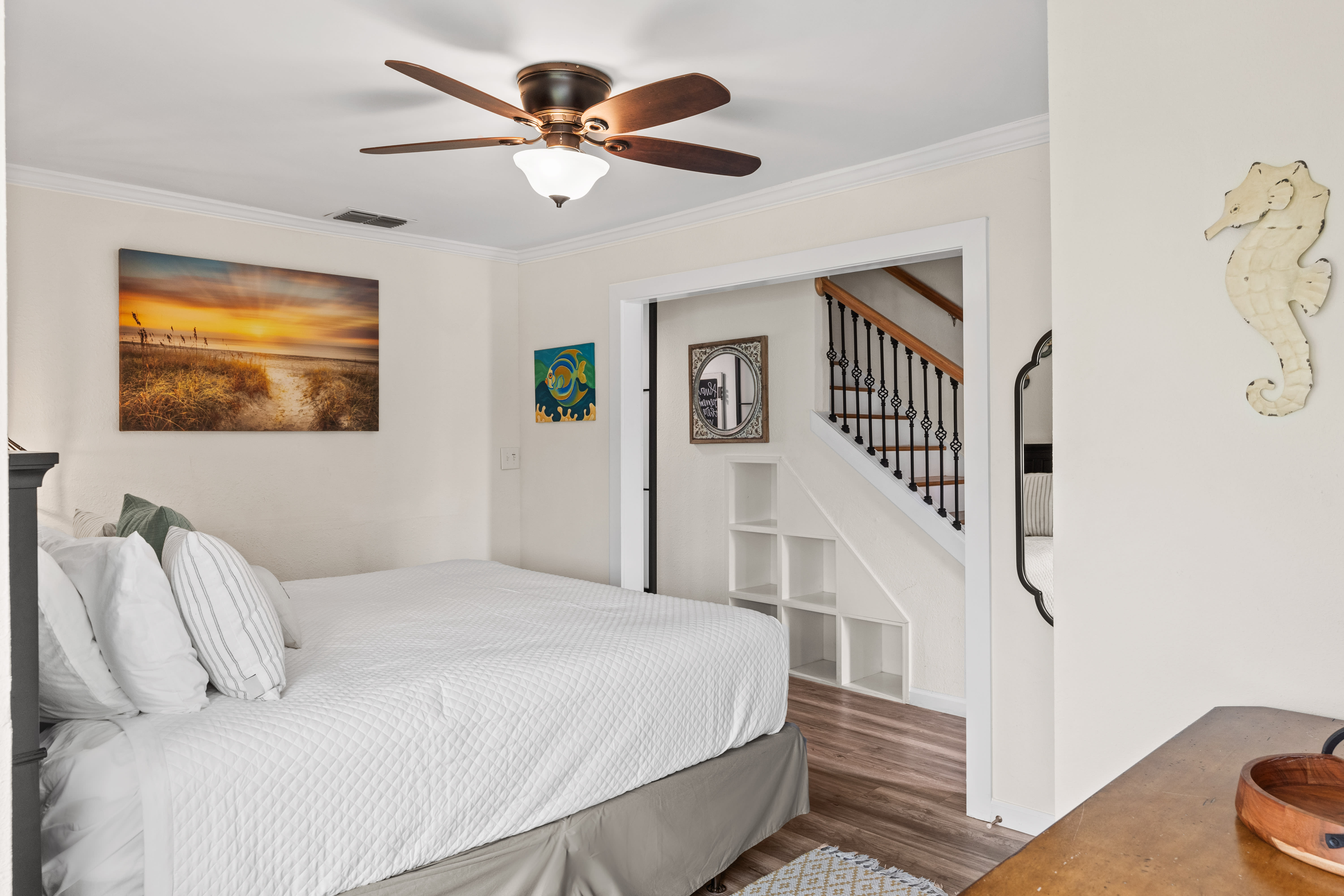 Unwind in your coastal-themed bedroom featuring crisp white linens, a cooling ceiling fan, and charming beach artwork that sets the perfect vacation mood.