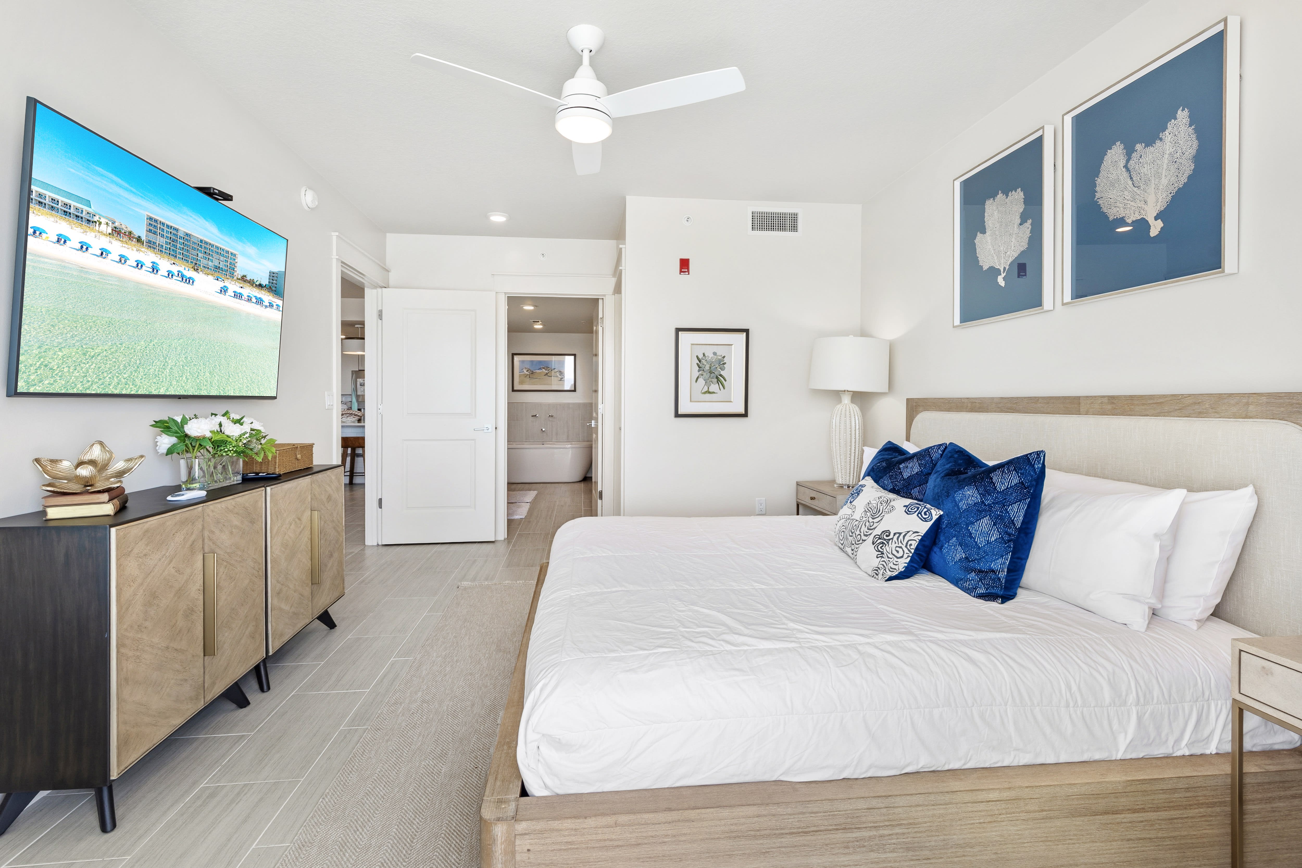 Unwind in your bright coastal bedroom featuring crisp linens, ocean-themed artwork, and a large TV for relaxation.