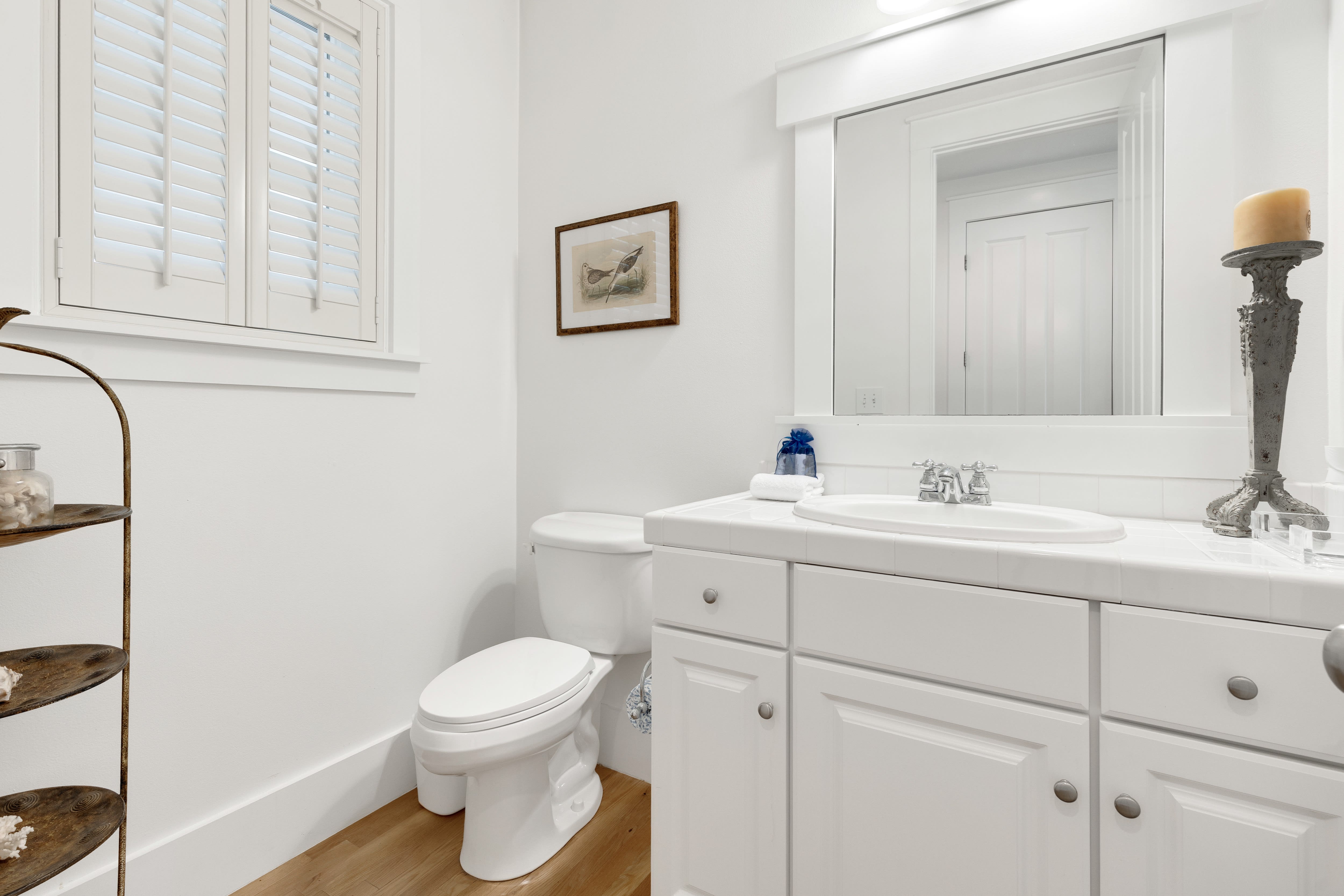 Your pristine white bathroom features warm wood floors, elegant shutters, and thoughtful touches like fluffy towels and coastal artwork.