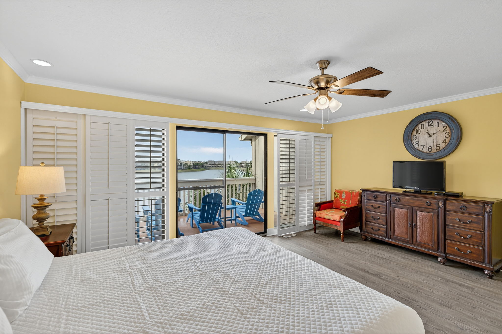 Wake up to waterfront views in your bright, comfortable bedroom with ceiling fan, TV, and private balcony access featuring blue Adirondack chairs.