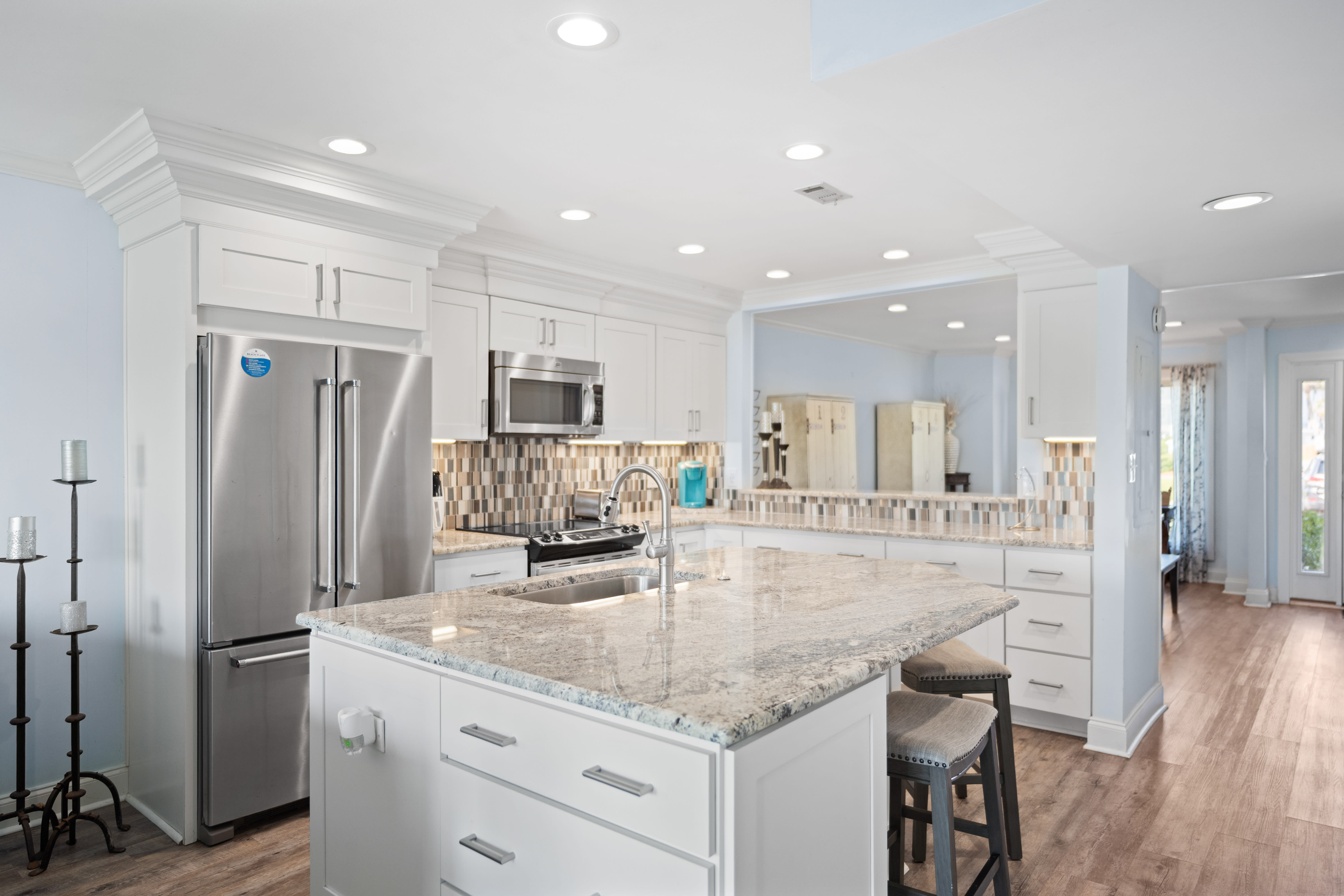 Modern kitchen with a large granite island, stainless steel appliances, white shaker cabinets, tile backsplash, and recessed lighting. Features include bar seating, hardwood floors, and a mirrored wall for added depth.