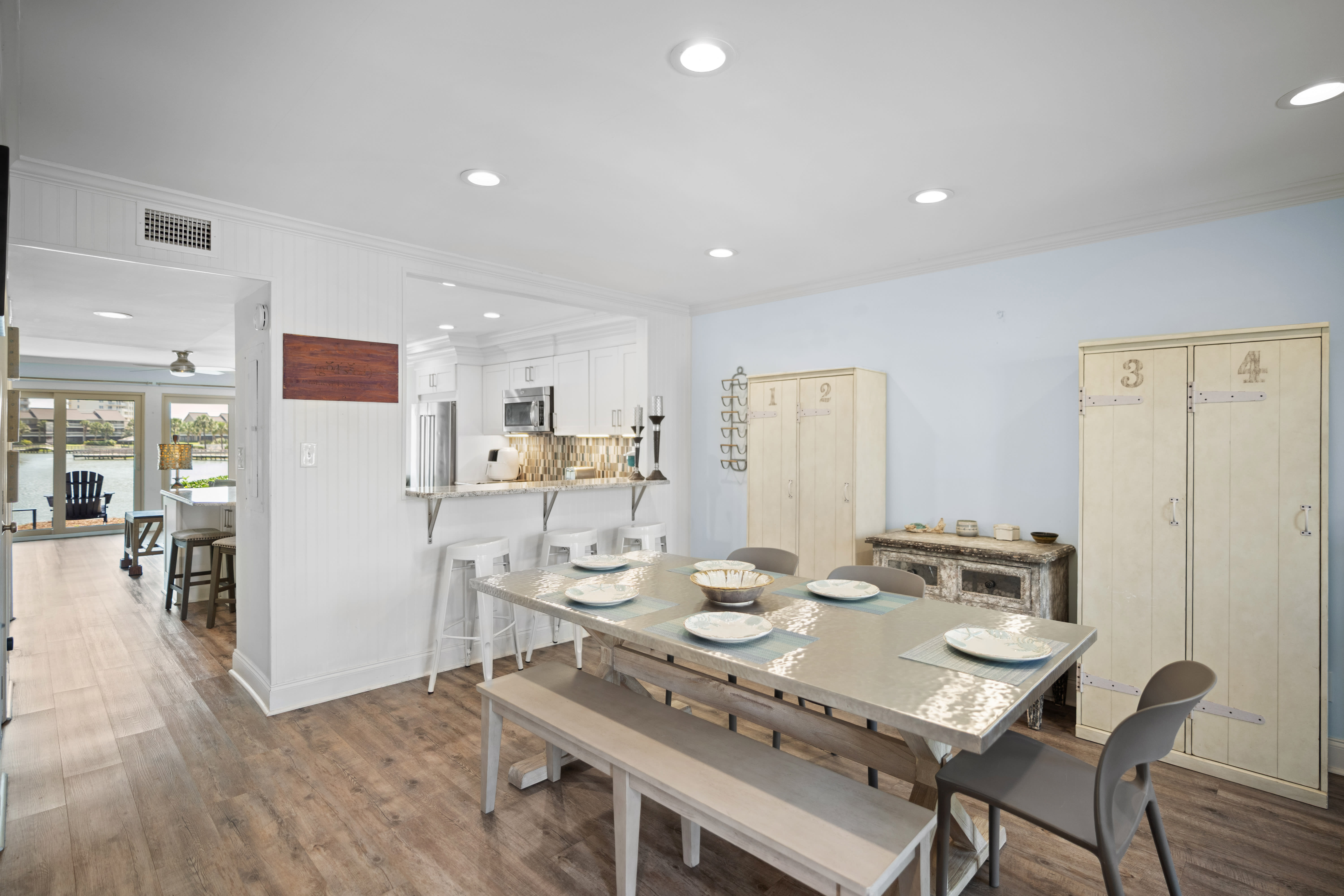Bright dining area with seating for six, barstools at the kitchen counter, and LVP wood floors. Features a clean coastal style with light furnishings and an open layout.