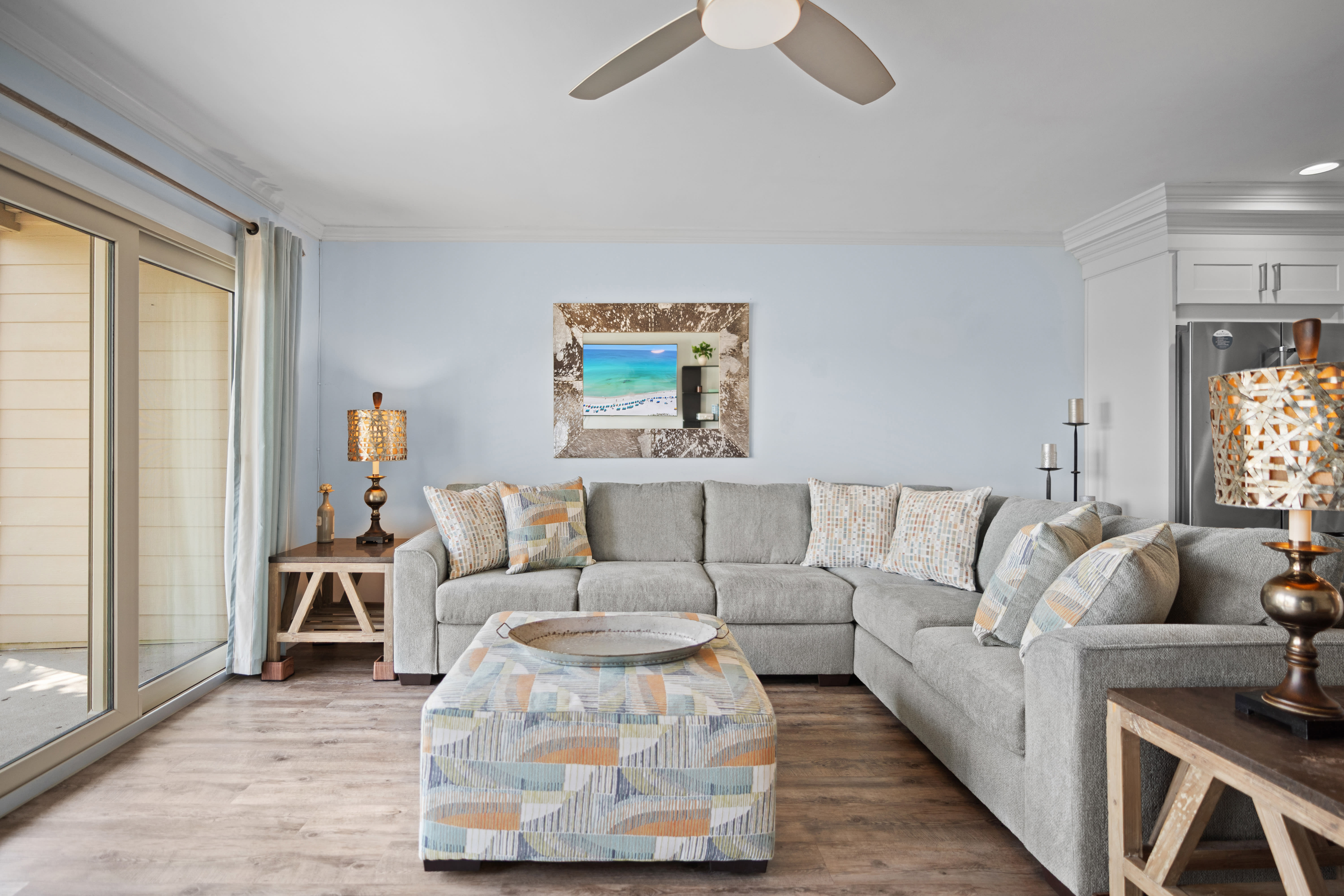 Cozy living room with a large sectional sofa, patterned ottoman, warm LVP wood floors, and beach-themed decor. Soft tones and natural light create a relaxed, inviting atmosphere.