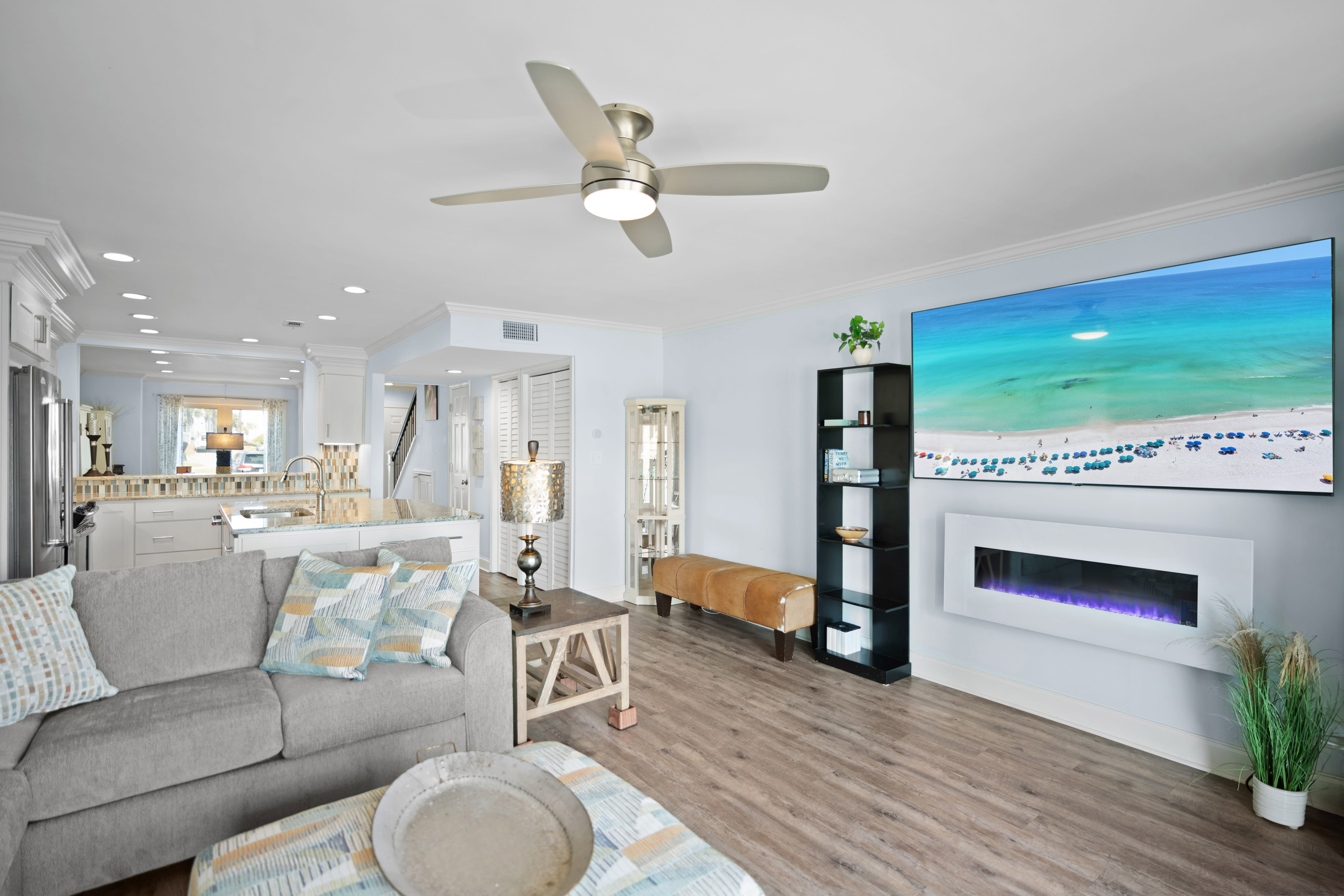 Open, coastal-style living space with LVP wood floors, large screen tv, modern electric fireplace, and beachy decor. Features comfy seating, ceiling fan, and a seamless flow into the dining and kitchen areas.