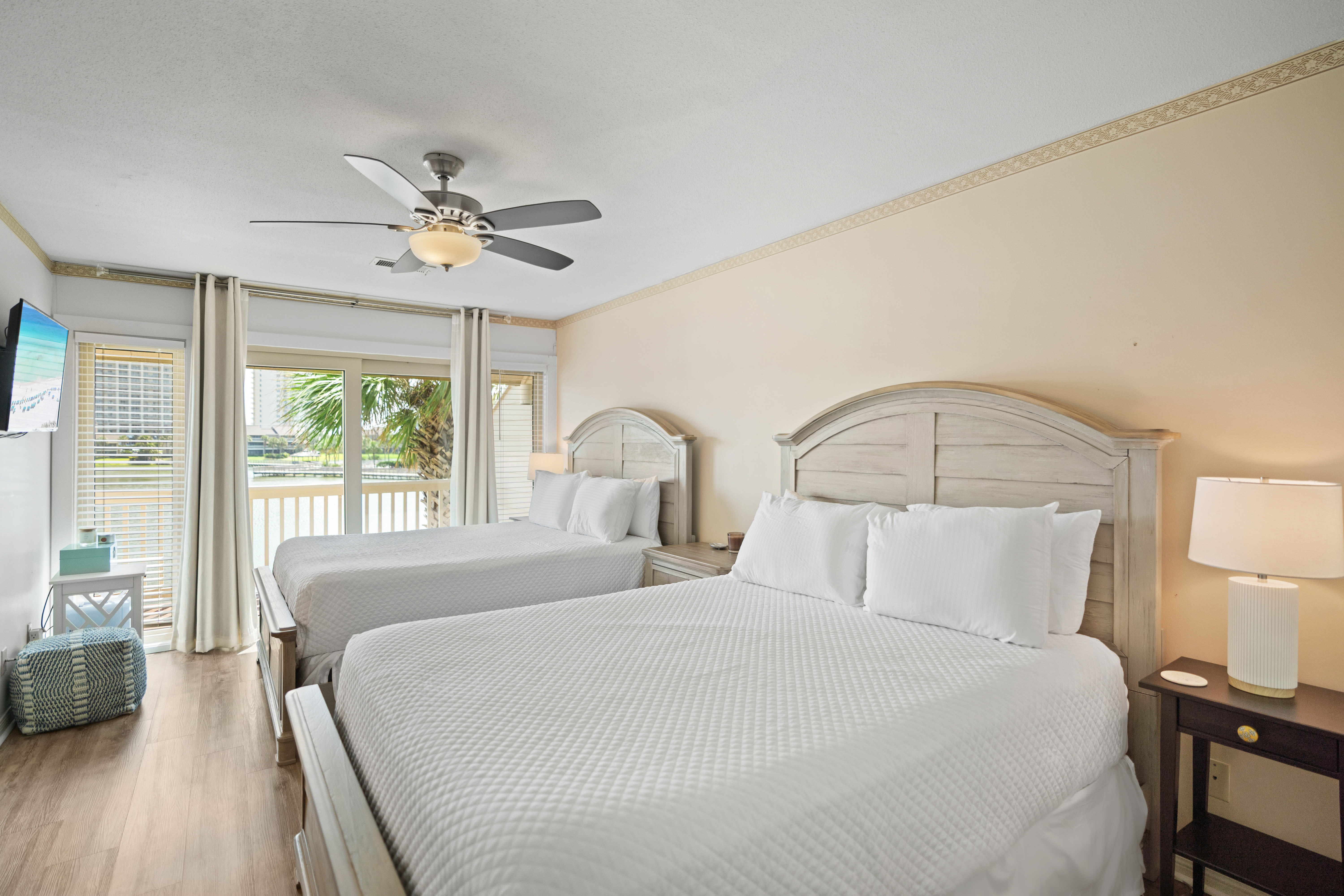 Bright guest bedroom with two queen beds, crisp white bedding, and light wood furniture. Features a ceiling fan, warm neutral walls, and access to a balcony with lake views.