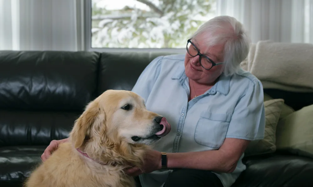 A woman named Marilyn is sitting on a couch in a living room, petting a friendly golden retriever dog that has its tongue out of its mouth.