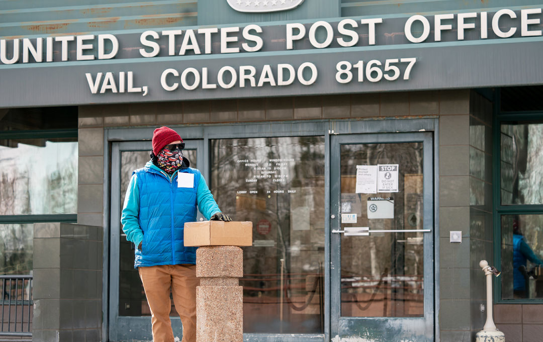 A person wearing a knit hat, balaclava, sunglasses, and gloves stands outside the Town of Vail Post Office with a package