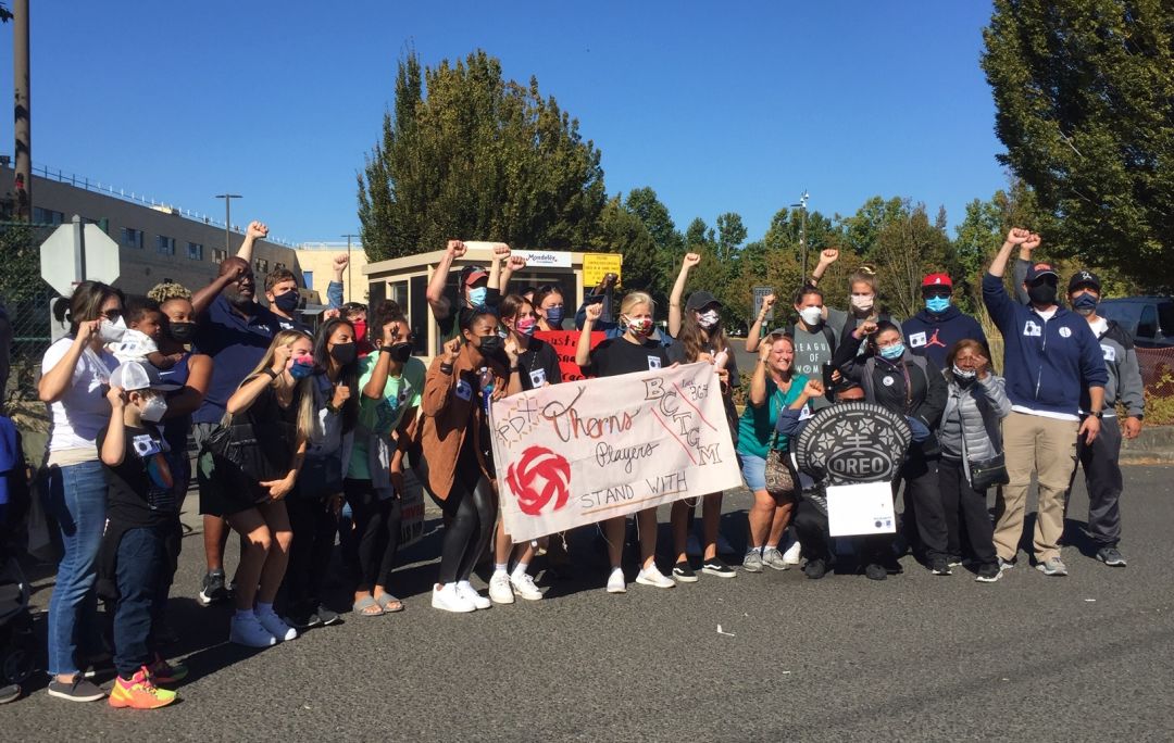 A group of people, one in an Oreo costume, with raised fists and a banner that says PDX Thorns players stand with BCTGM Local 364