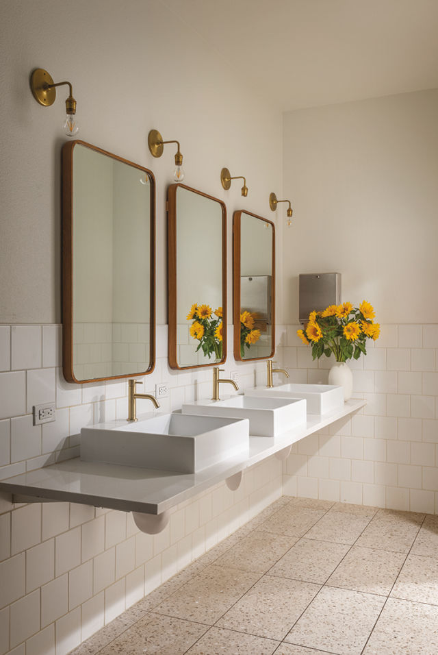 a row of three bathroom sinks and mirrors, with a vase of yellow flowers on the counter