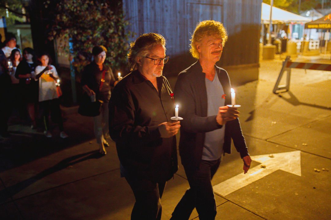 Two masculine-presenting people holding candles and walking.
