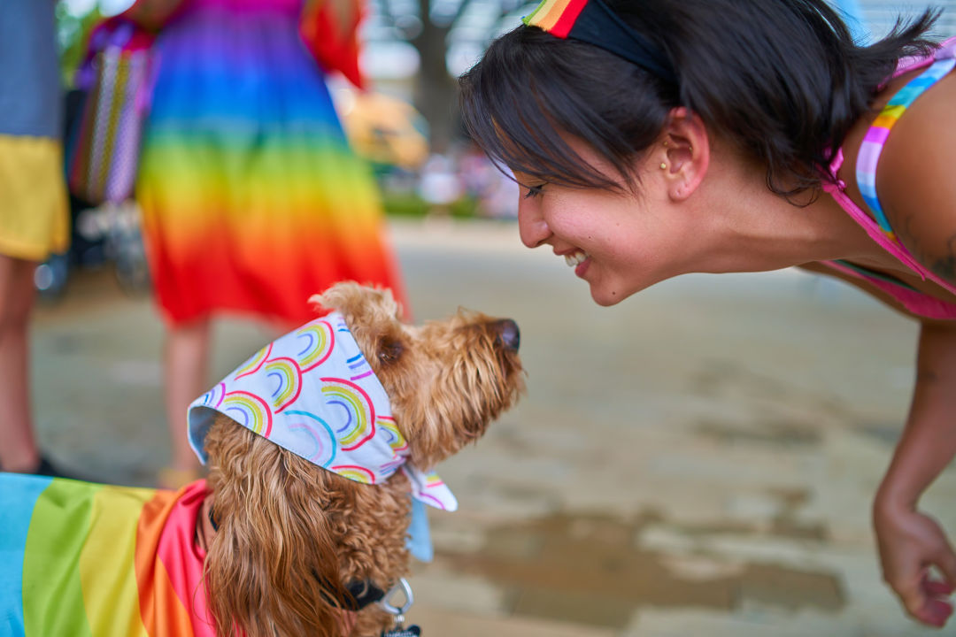 A feminine presenting person bends over and smiles at a dog in a rainbow bandana.