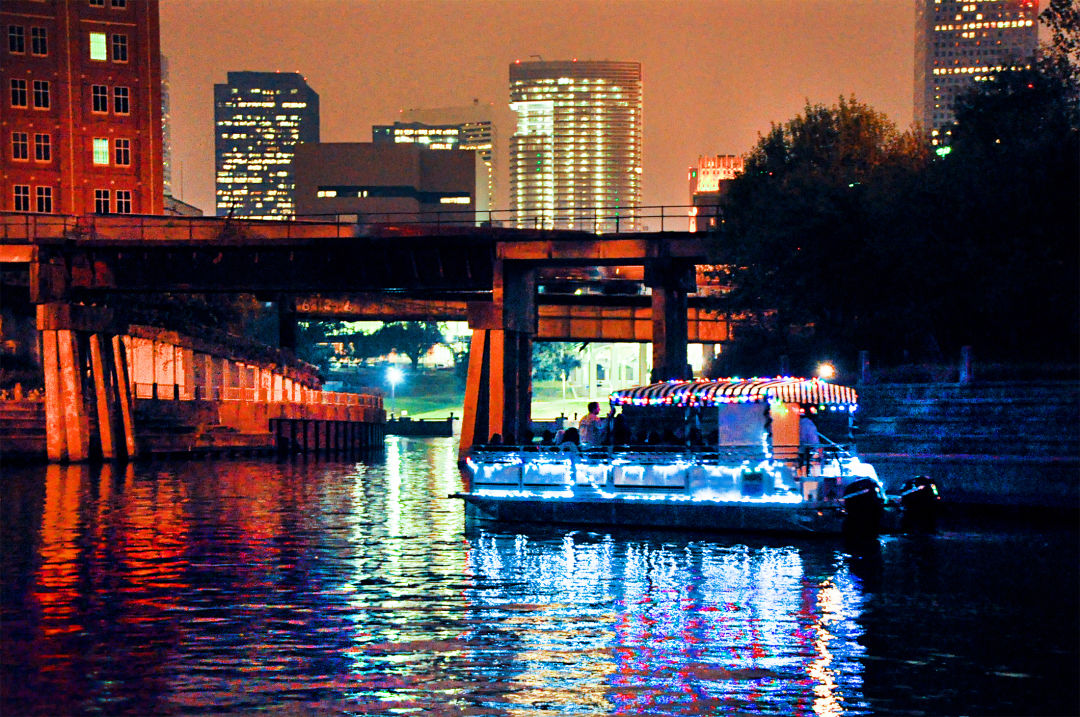 A pontoon boat lit up for the holidays. It floats on the bayou in front of the downtown Houston skyline.