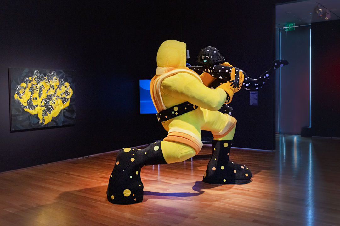 Soft sculpture of a space man in a black and yellow suit and a sentient star in the shape of a woman embracing. There is a painting of them behind them.