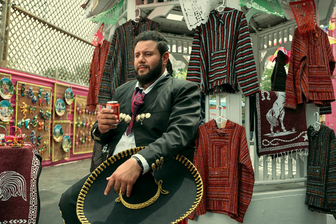 A man drinks a can of Coca-Cola while dressed as a mariachi and sitting in front of a market stall selling Mexican crafts.