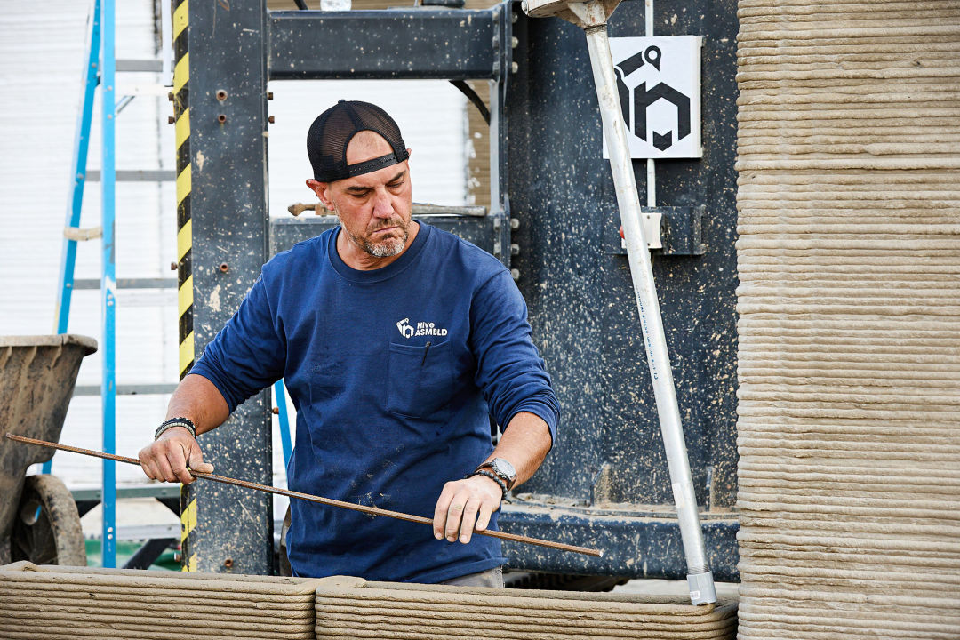 Osoba prezentująca męskość, ubrana w niebieską koszulę i czarną czapkę z daszkiem skierowaną do tyłu, stoi przy drukarce 3D, która drukuje beton.