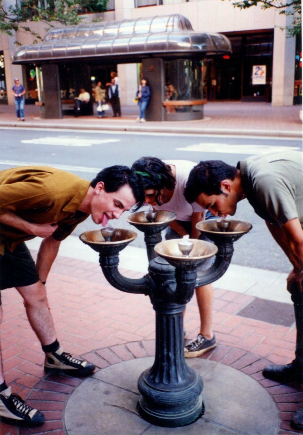 Three people lean over a Benson Bubbler water fountain on a downtown street with a bus shelter behind them