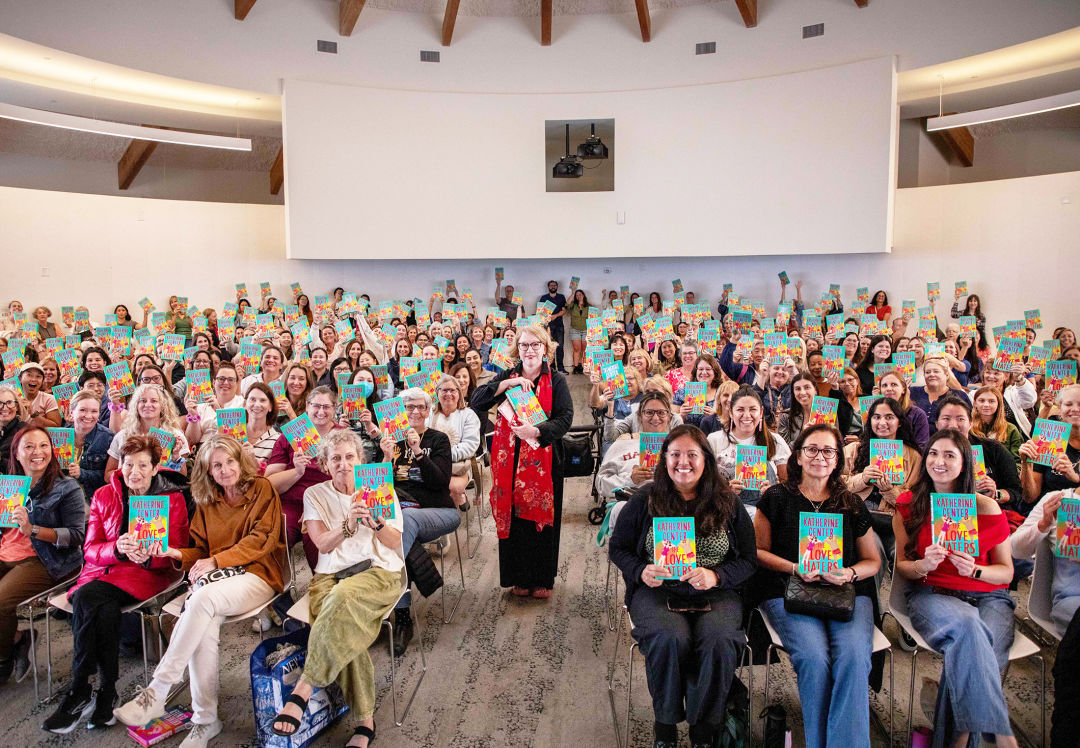 A large audience holding up copies of a book while the author stands in the middle doing the same.