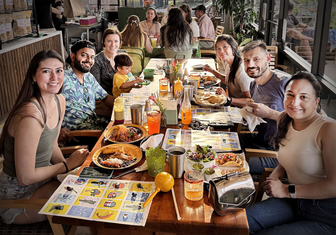A group of people smiling at the camera while they play lotería and eat Mexican food.