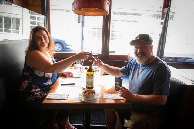 Two people clinking wine glasses at a restaurant table with a McMenamins passport