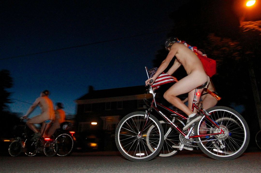 A naked person seen in profile riding a bicycle at night, with other naked people on bikes in the background