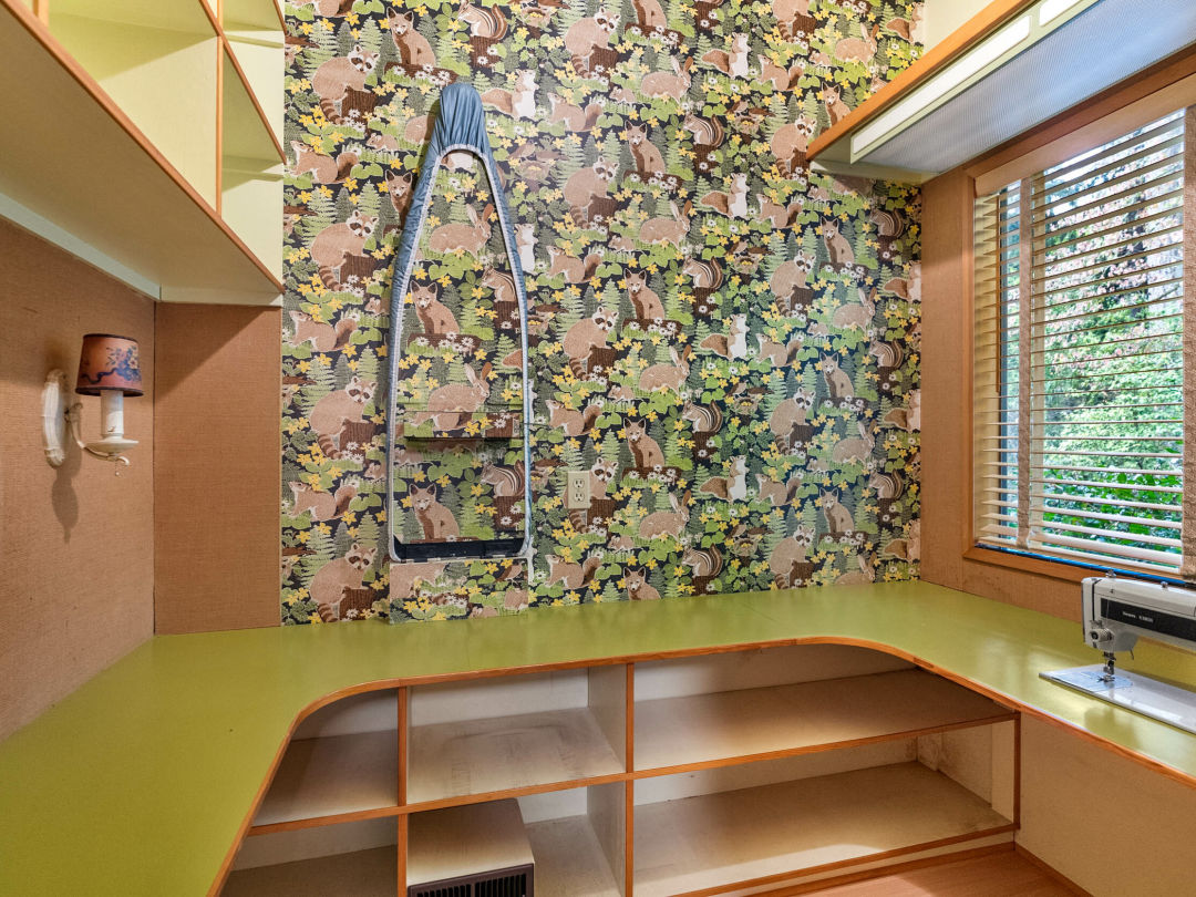 Room interior with shelving, rounded counter, sewing machine, and fox and raccoon printed wallpaper with a folded up ironing board camouflaged in the wallpaper