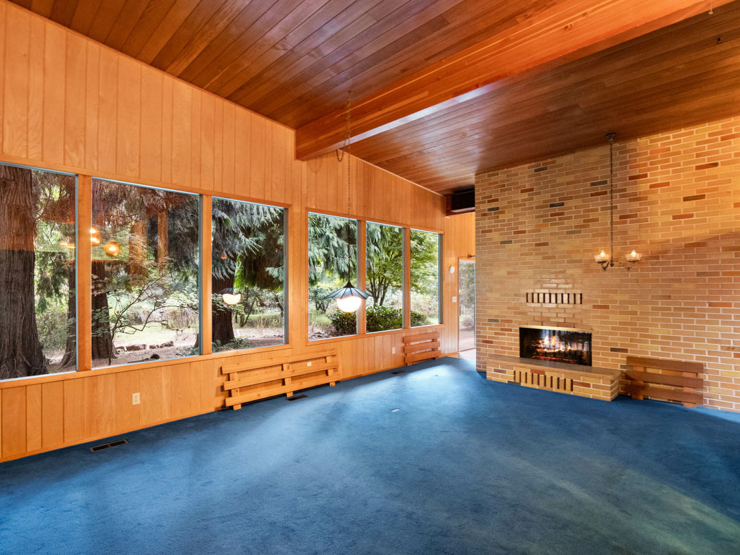 A room with blue carpet, lots of wood paneling, no furniture, a fireplace in a brick wall, and a view of trees outside large windows