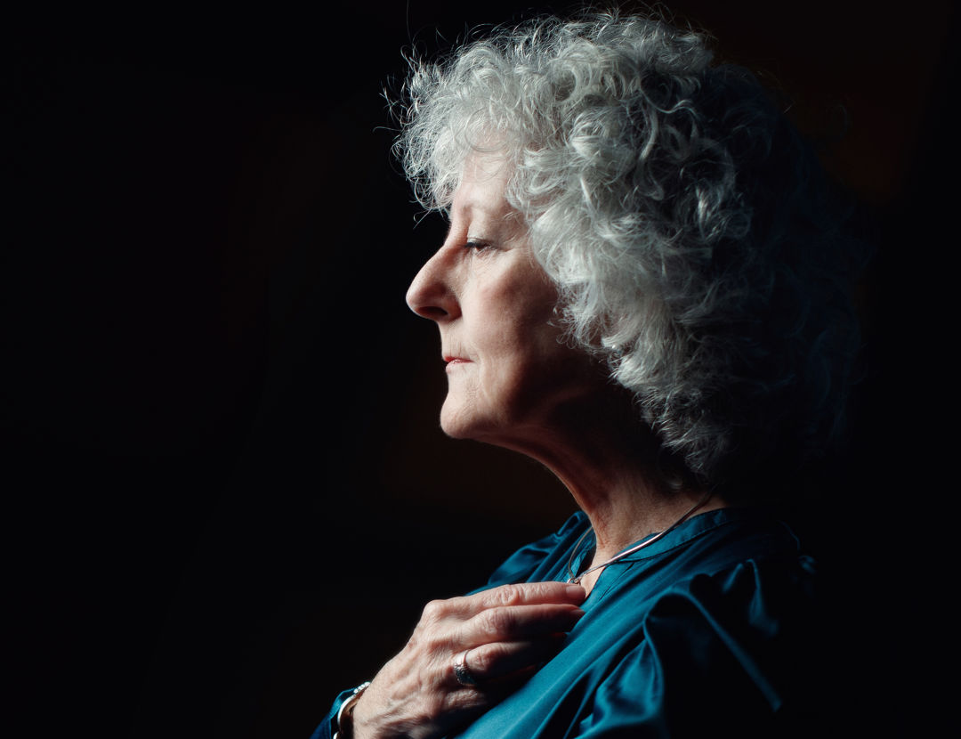 A grey-haired woman clutches her hand to her chest and looks to the left.