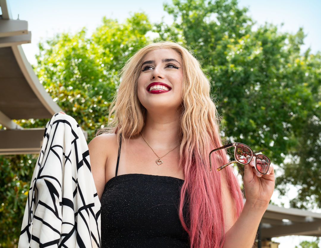 A blonde femme-presenting person holds up a pair of sunglasses and a blazer while looking up and smiling.