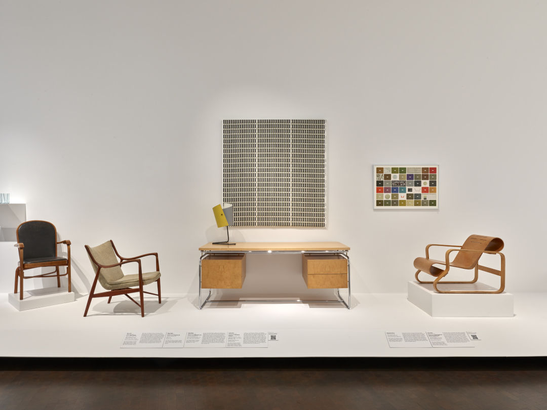 A midcentury modern desk in a museum exhibition. A lamp sits on it and an abstract expressionist art piece hangs behind it. There are chairs next to it, each with a different design.