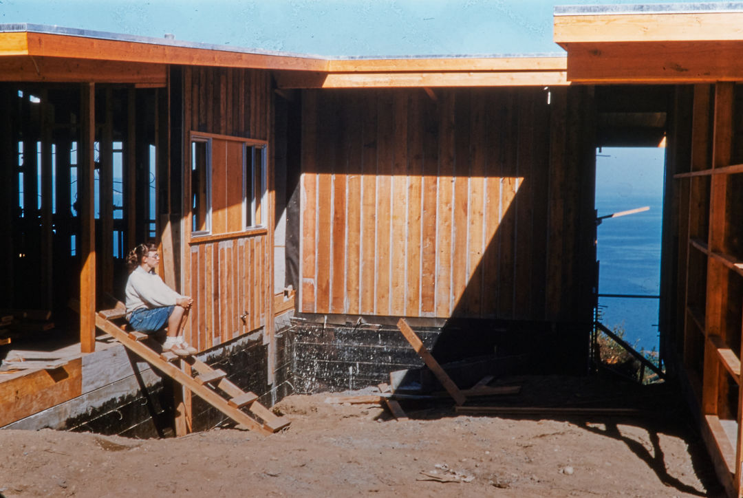 A 1950s photograph of a woman sitting in an unfinished house's timber framing