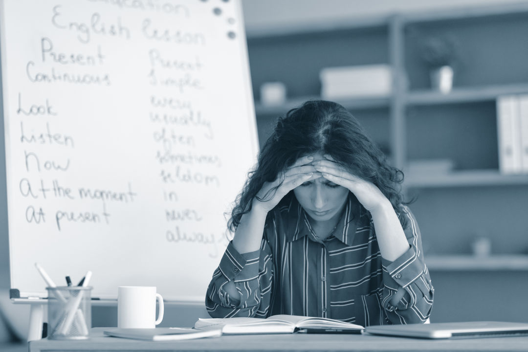 A stock image of a teacher at her desk holding her head in frustration.