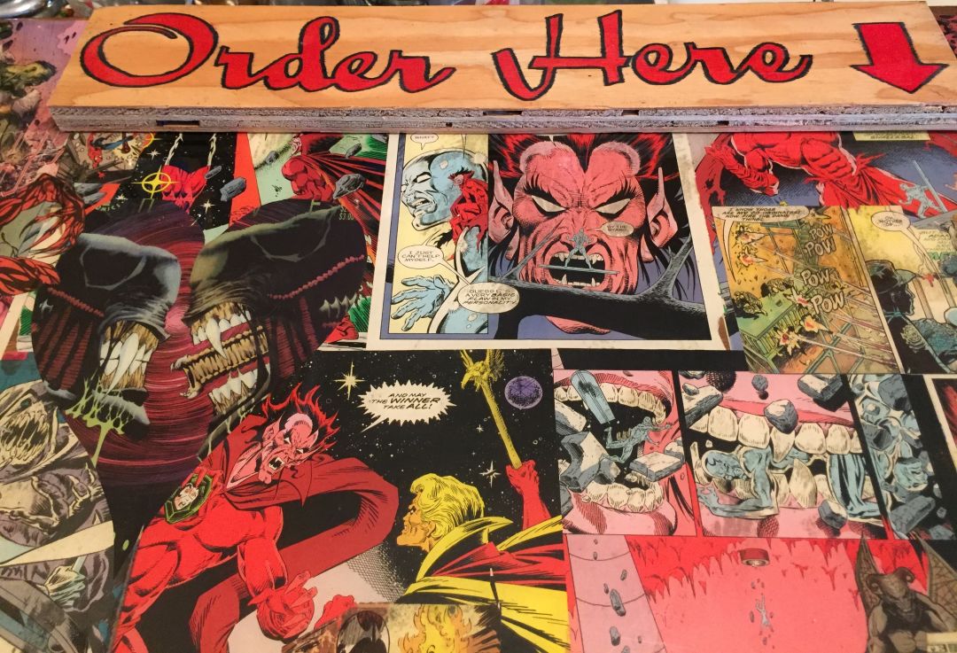 An "Order Here"sign with an arrow, affixed to a surface covered in comic book images