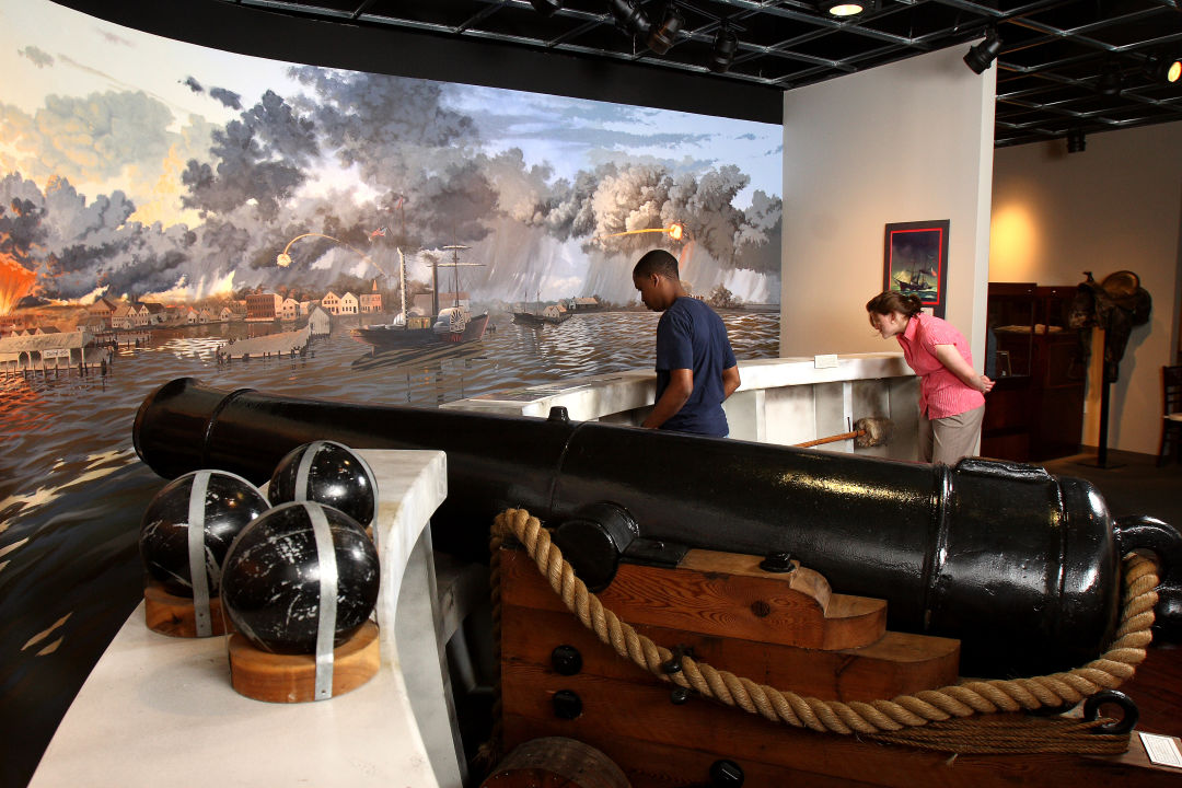 Two people browse a museum exhibit with cannons and a war scene.