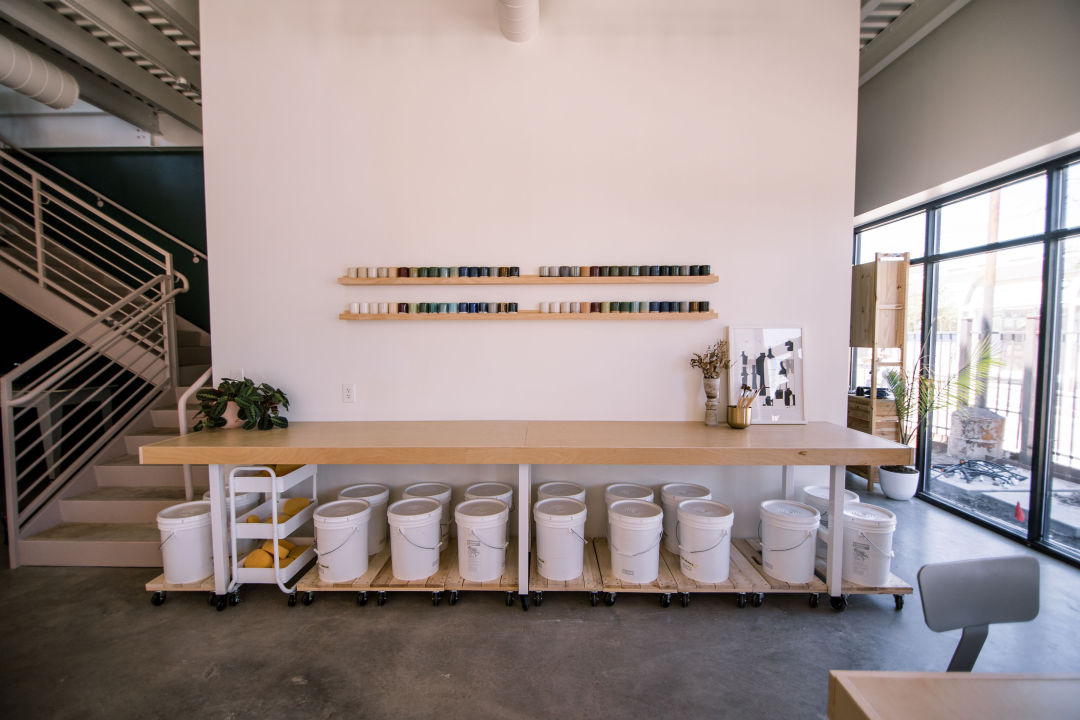 A wall of clay options, including samples and buckets, sit on, are hung on a wall, and are stored below a table. There is a stairwell behind the wall.