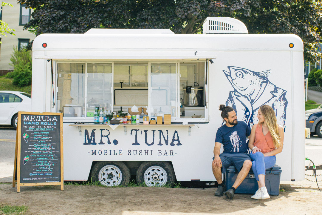 A smiling man and woman sitting in front of Mr.  Tuna mobile sushi bar