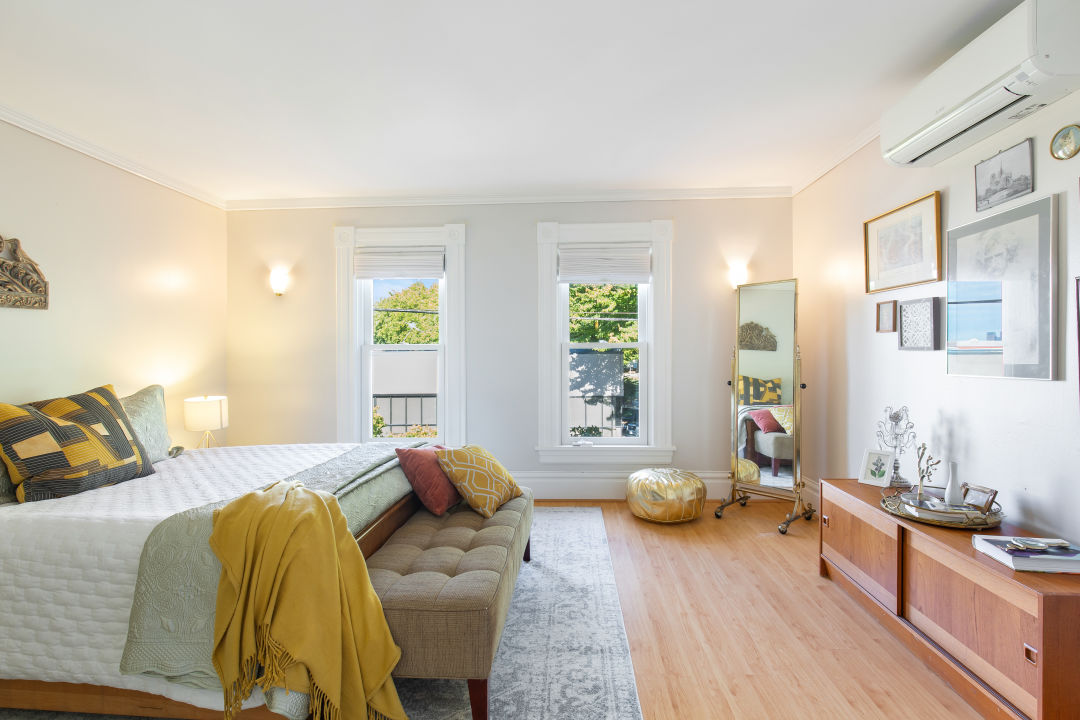 A bedroom with two white-trimmed windows on the far wall, a bed with its head on the left wall, and a low cabinet with framed photos above on the right.