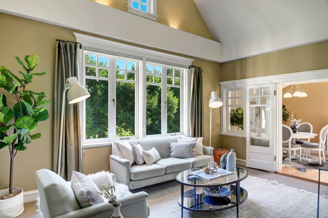 A living room with mustard walls and white trim. A three-window bank is surrounded by decorative moulding. In the back, a glass door and window peek into a dining room.