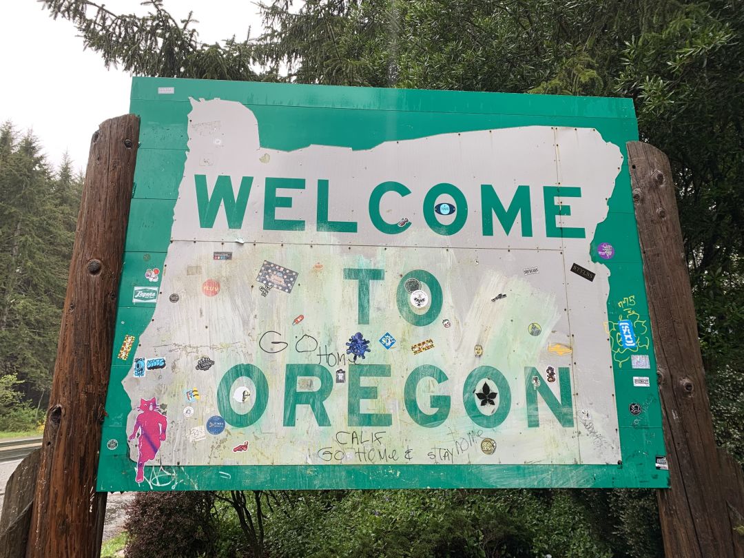 Green "Welcome to Oregon" highway sign with many stickers and scrawled message, including "Go Home" and "Calif Go Home & Stay Home"